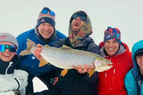 Five people smiling, one holding a large fish, all wearing winter clothing.