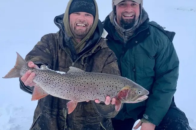 Two men in winter clothing smiling with a large fish in snowy landscape.
