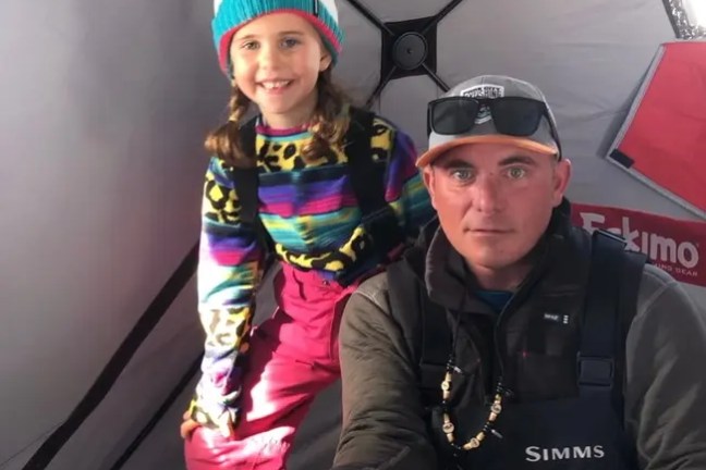 Man holding fish, child smiling beside him in ice fishing tent.